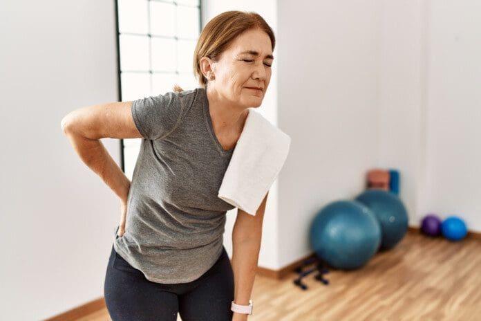 Middle age woman wearing sporty look training at the gym room suffering of backache, touching back with hand, muscular pain