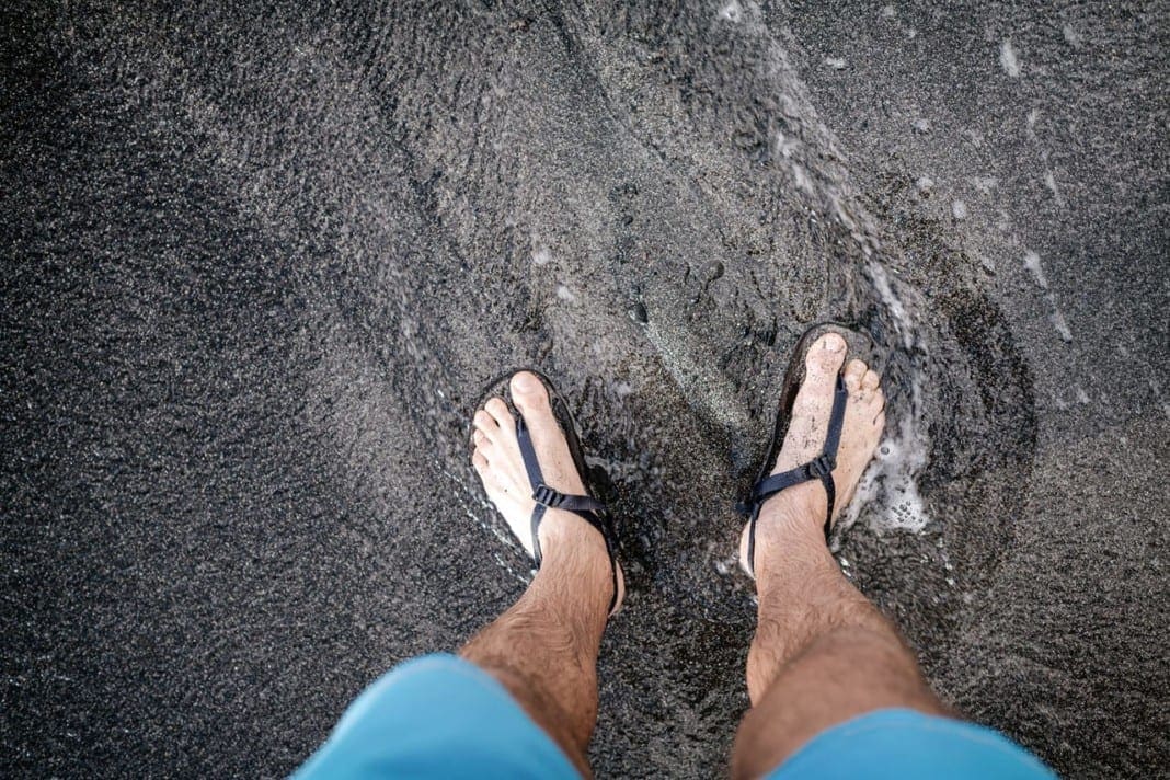 Man looking down at feet and sandals on volcanic black sand beac 11860 Vista Del Sol, Ste. 128 Summer Footwear, Back Pain and What To Know