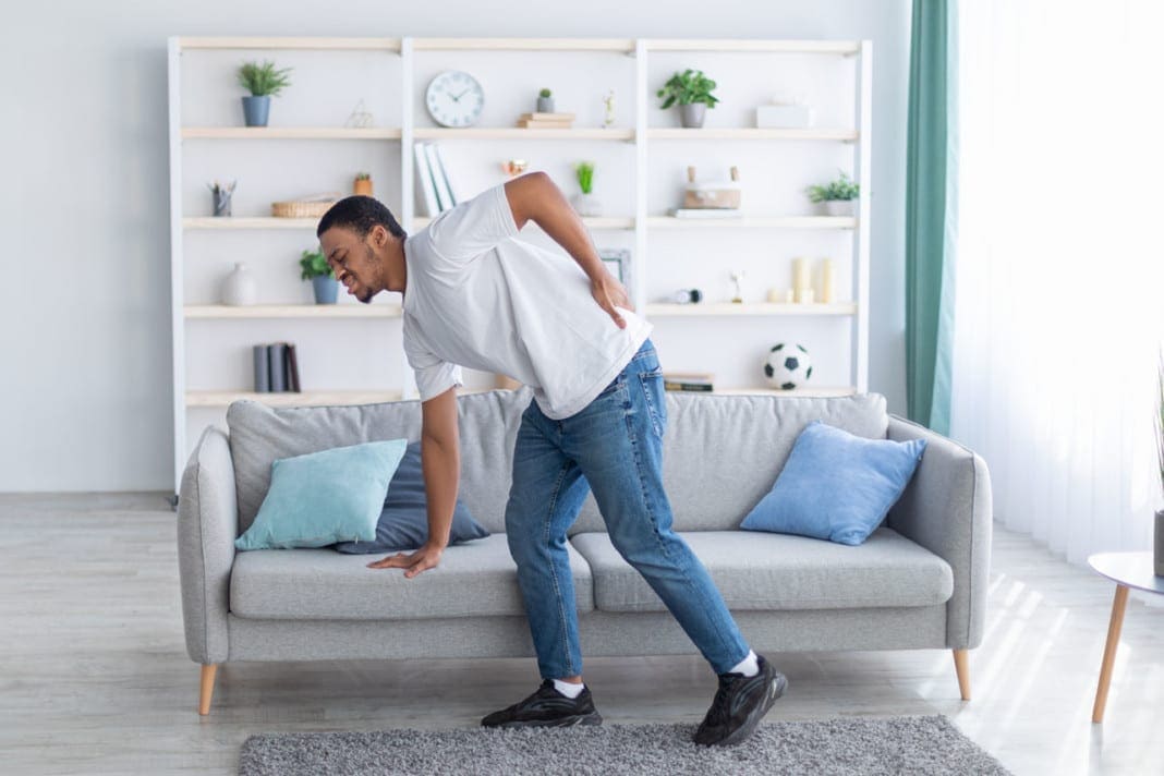 Millennial African American guy suffering from back pain, leaning on sofa, cannot walk at home. Healthcare concept