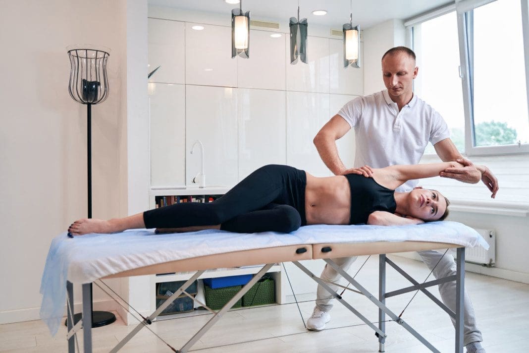 Handsome male physiotherapist in white shirt stretching woman back on massage table in medical office