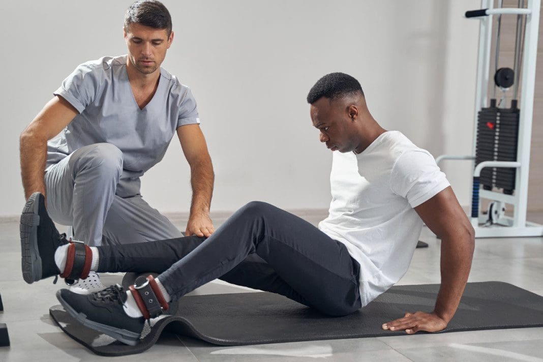 Concentrated man foam-rolling his calf muscles supervised by physiotherapist