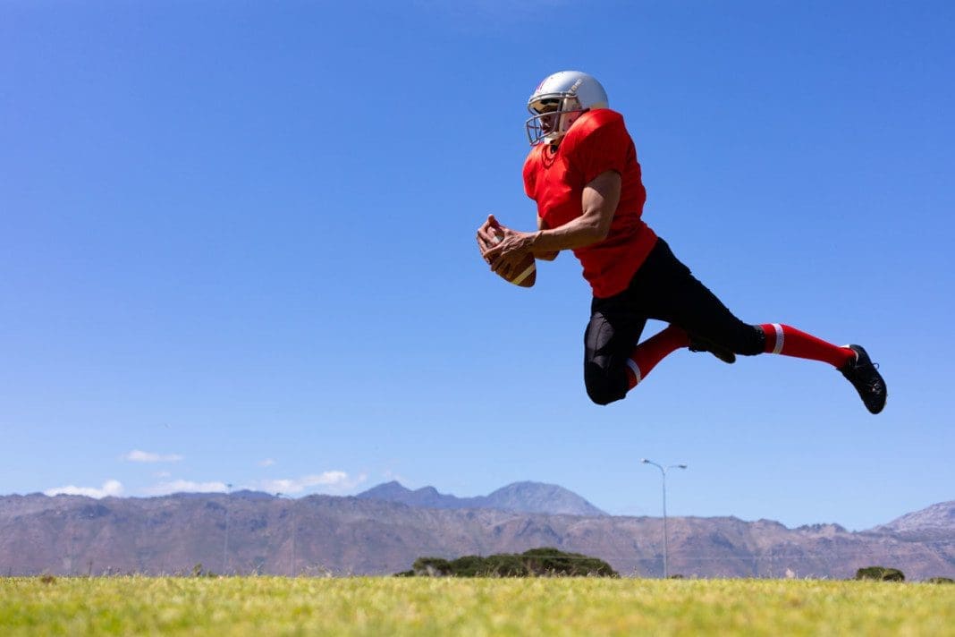 Side view of a mixed race male American football player wearing a team uniform, training at a sports field, jumping in the air holding a football, with blue sky in the background