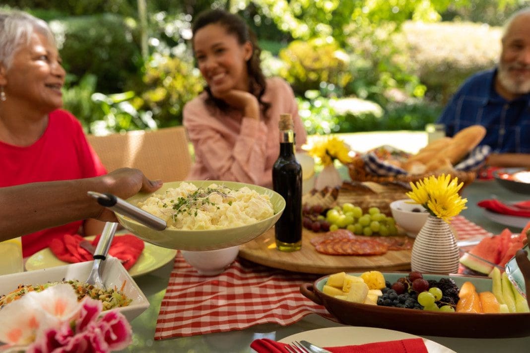 Side view of a multi-ethnic adult family serving food sitting at a table, talking and smiling during a meal together outside on a patio in the sun
