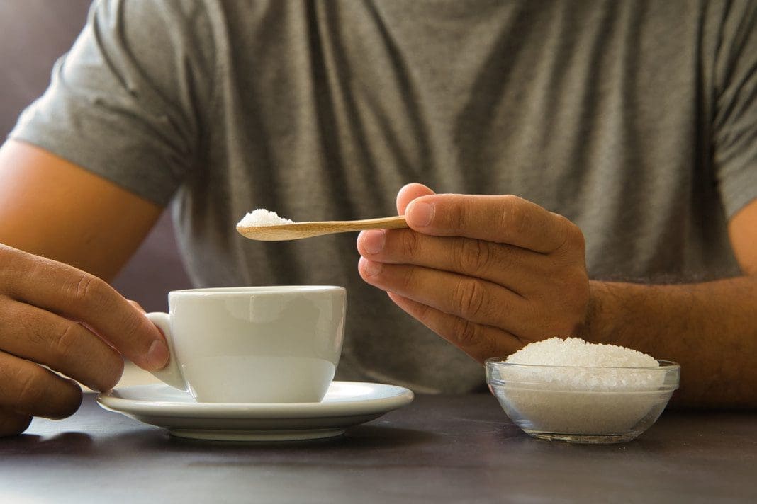 Male hand holding wooden spoon with sugar over cup of coffee or