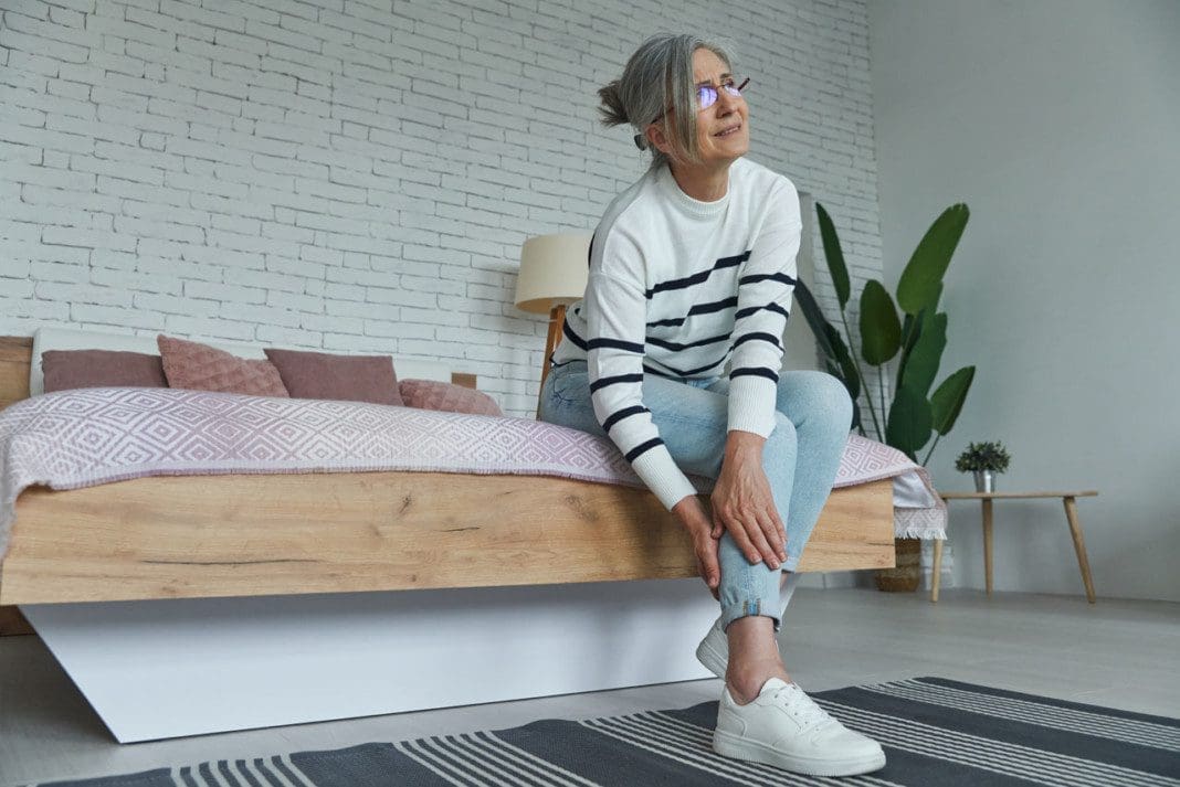 Senior woman suffering from leg pain while sitting on the bed at