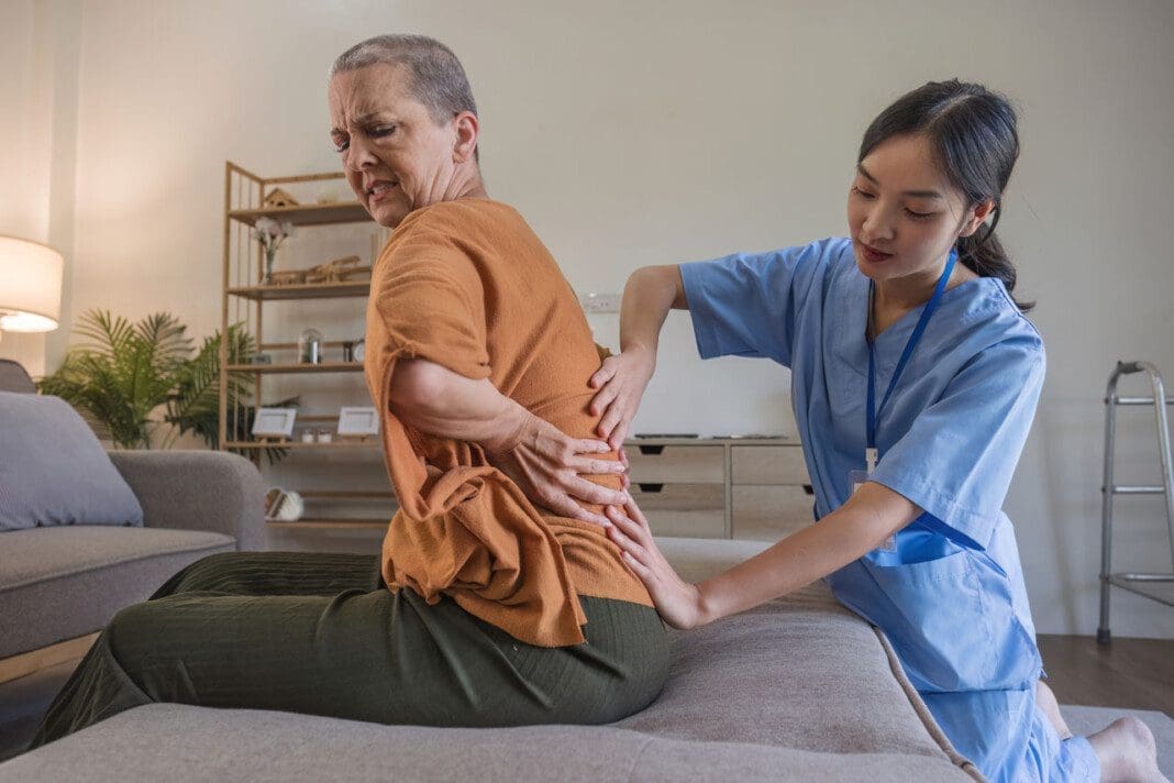 Caucasian senior woman 60s doing physiotherapist with support from nurse. Senior elderly female sitting on sofa in living room workout exercise for patient with caregiver in nursing care.