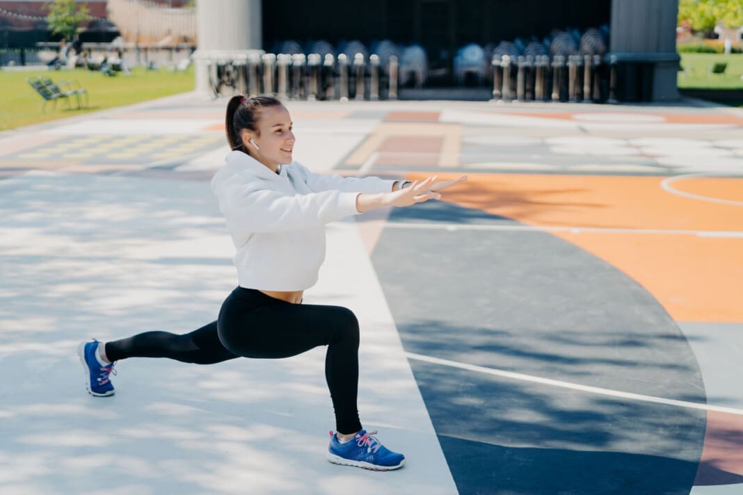 Horizontal shot of motivated sporty woman stretches legs and arms does warming up exercises before workout looks forward dressed in sportsclothes poses outside on big stadium. Exercising concept